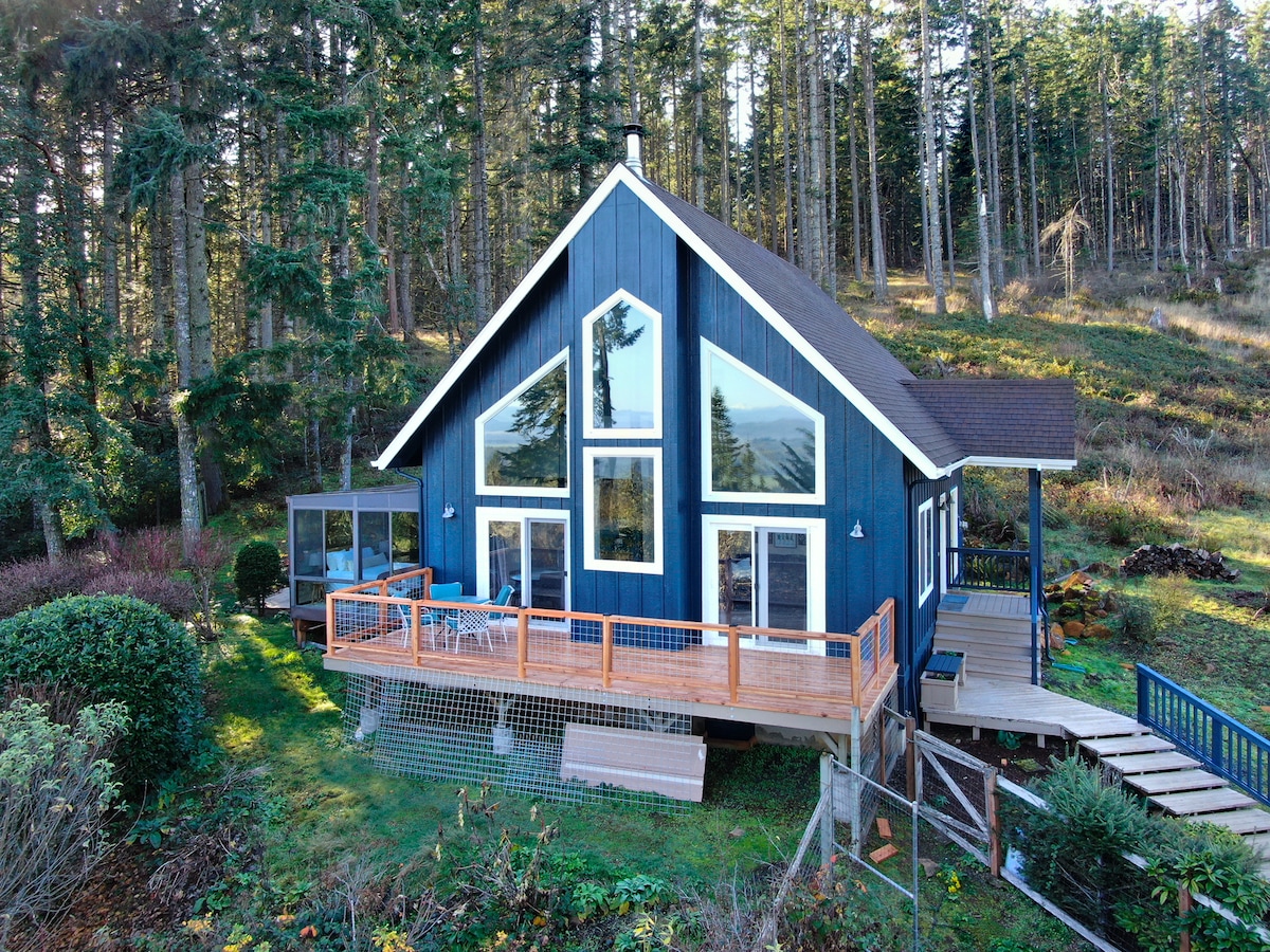 The exterior of the chalet-style house showcases a striking navy blue facade with large windows that offer views of the surrounding trees. A spacious deck extends from the house, complemented by a path leading to the entryway. Natural greenery surrounds the property.