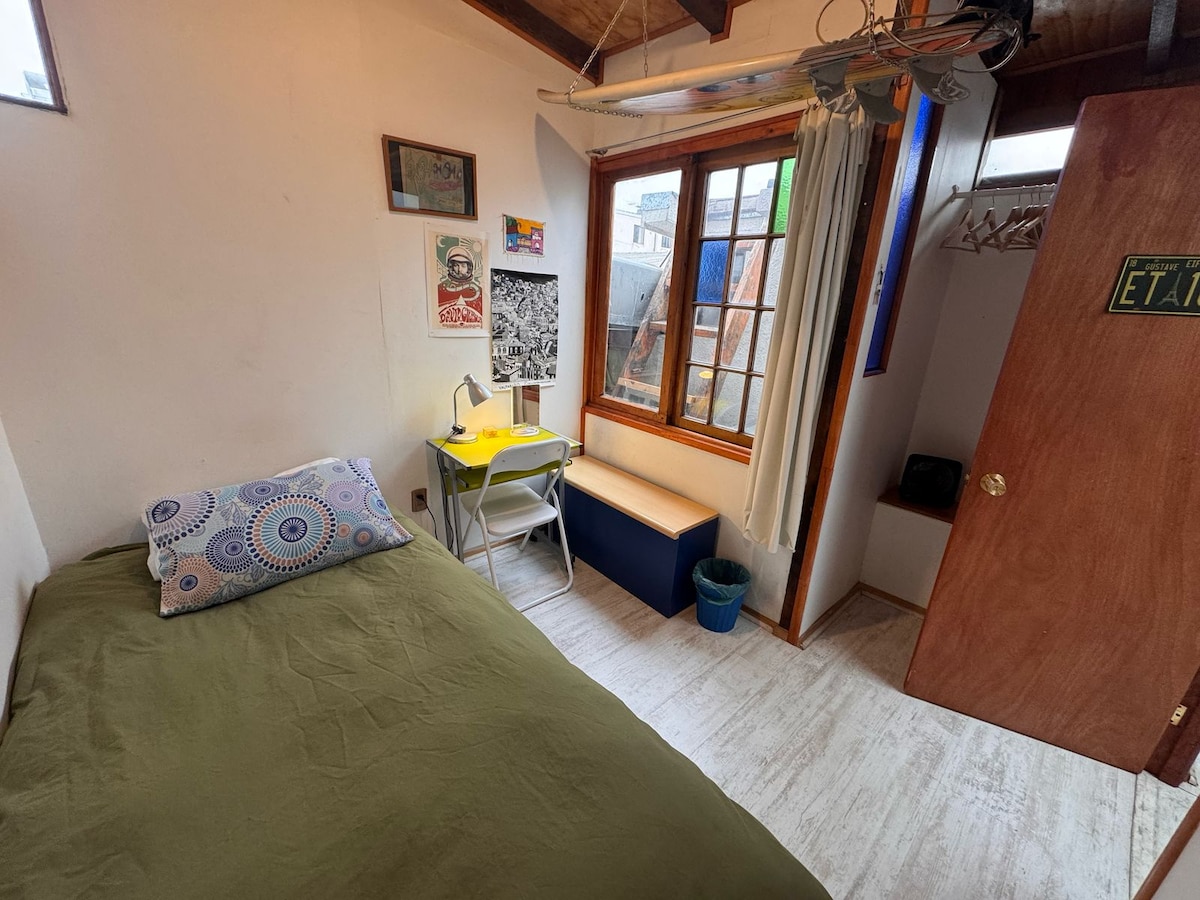 A well-lit room features a spacious bed with green bedding and decorative pillows. A small desk with a lamp and chair is positioned beside a window, which offers natural light and views. A closet area is visible, along with a wooden door leading outside.