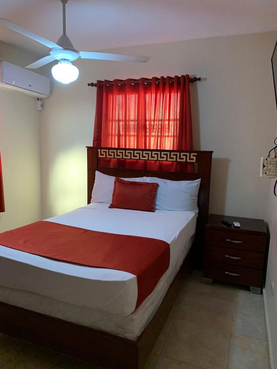 A cozy sleeping area is presented with a wooden bed featuring a decorative headboard. Crisp white linens and a red accent pillow are arranged on the bed. A dresser with multiple drawers is positioned nearby, while a ceiling fan and large window with red curtains offer light and ventilation.