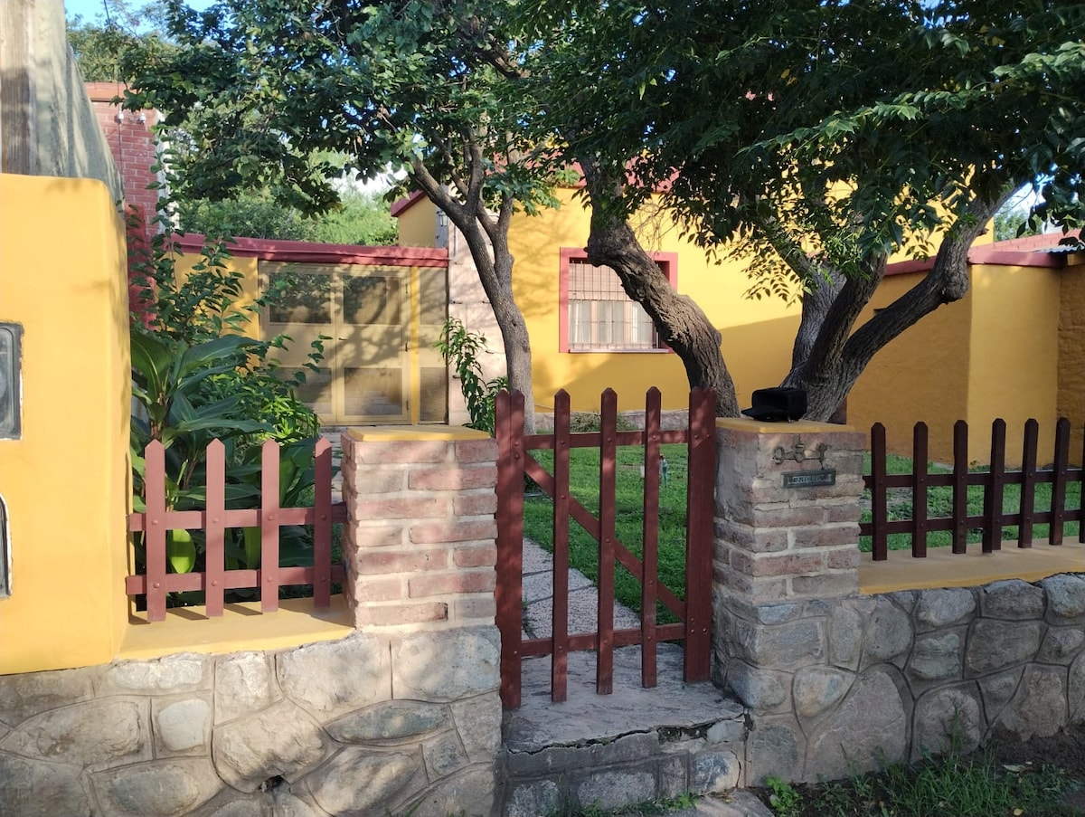 A charming entrance showcases a picket fence made of brown wood, leading to a small stone path surrounded by green grass. Lush trees provide shade, and a brightly painted yellow house is visible in the background. The welcoming setting emphasizes privacy and tranquility.