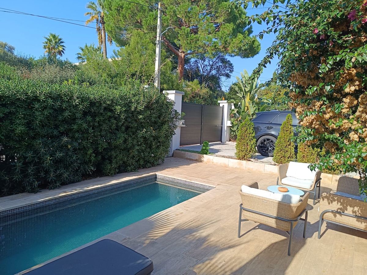 The image presents a private pool area surrounded by lush greenery and a well-maintained garden. The pool is set against a backdrop of palm trees, and lounge chairs are arranged nearby on a sunlit terrace. A car is parked in the driveway, and a gated entrance can be seen.