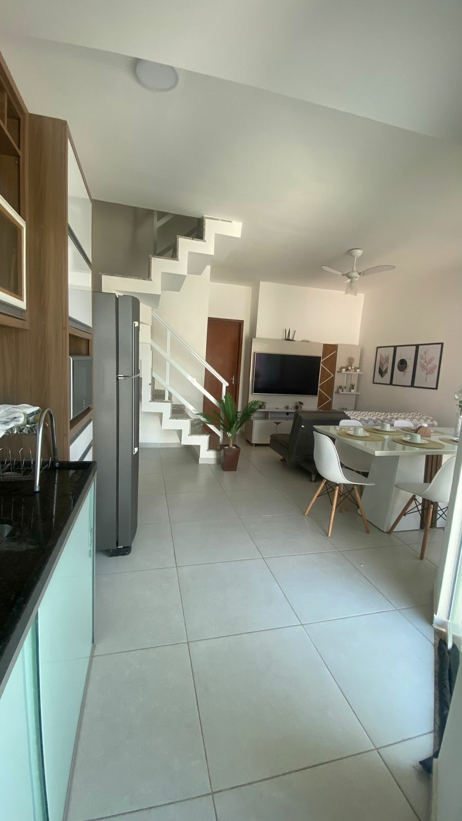An open-plan area features a modern kitchen with dark countertops and light cabinetry, leading to a dining space with a round table and several chairs. A staircase ascends in the back, and a wall-mounted TV is positioned nearby, enhancing the functional design.