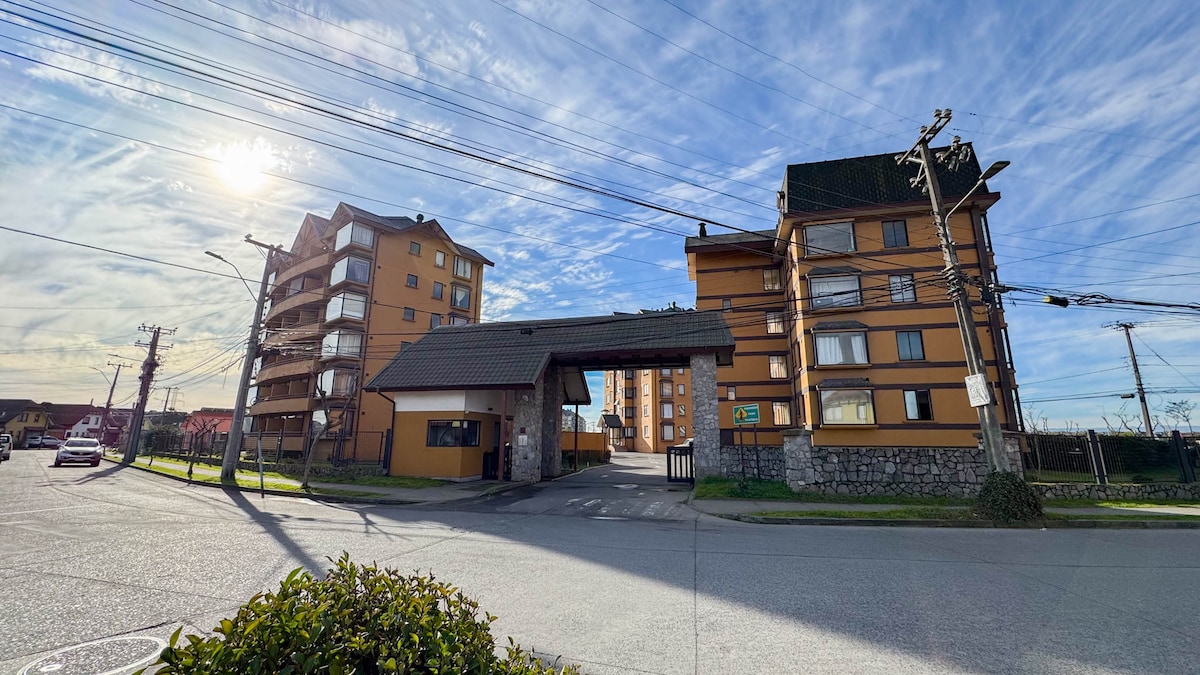 The entrance to a community of three apartment buildings is visible, showcasing a welcoming gate and well-maintained landscaping. The buildings exhibit a warm color palette, complemented by a clear blue sky with scattered clouds, indicating a pleasant day.