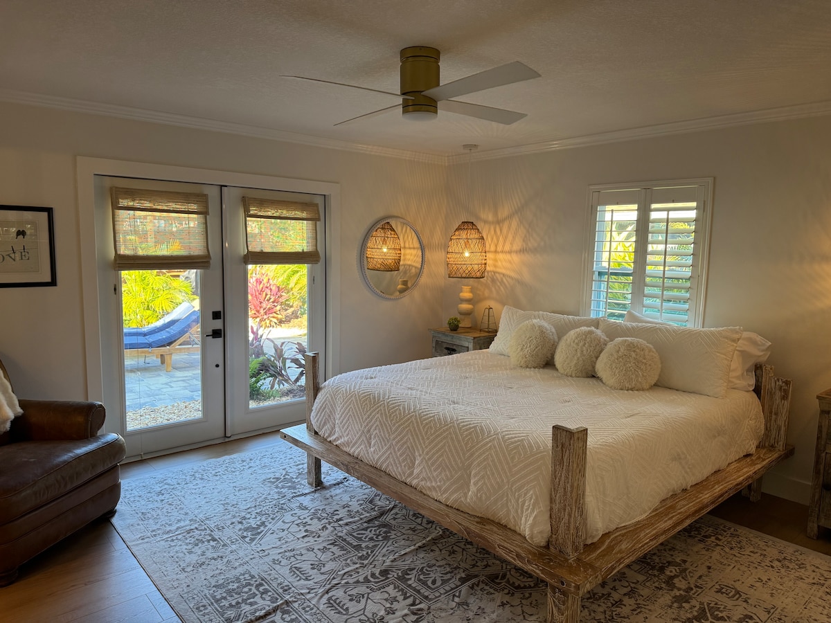 A spacious bedroom is furnished with a large bed featuring a textured white coverlet. Natural light enters through double doors leading to a garden, while two decorative mirrors and woven light fixtures add character to the room. A cozy seating area is present with a leather chair.