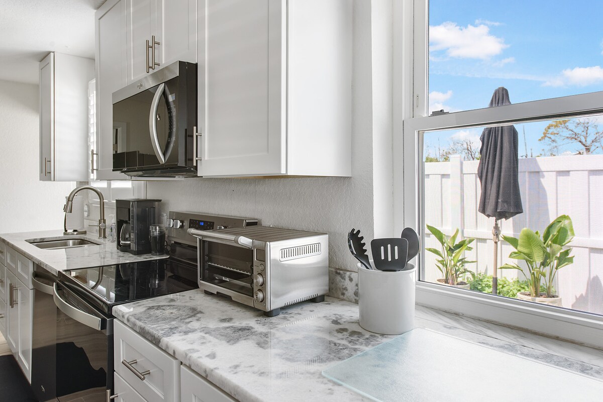 A modern kitchen with sleek white cabinetry and a marble countertop is depicted. Essential appliances, including a coffee maker, toaster oven, and microwave, are visible. Natural light streams in through a window, highlighting plants outside and enhancing the fresh, clean look of the space.