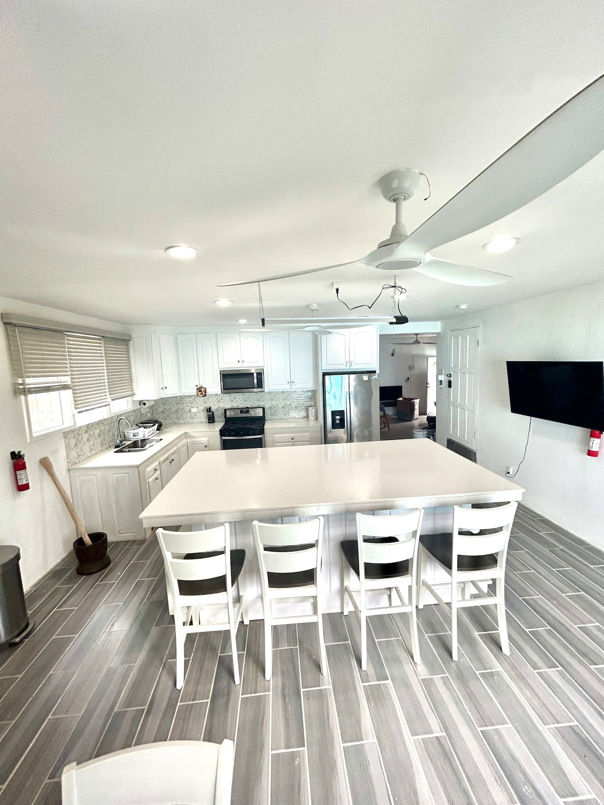The kitchen showcases white cabinetry and modern appliances, including a refrigerator and stovetop. A spacious island with seating for four is centered in the room. Natural light is provided by several windows, with sleek gray tiles covering the floor.