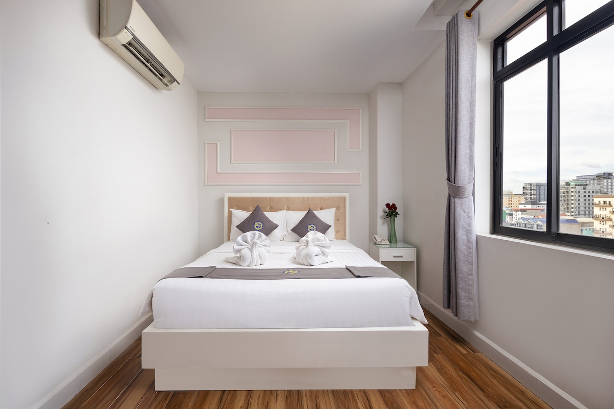 A bright guest room features a spacious bed with neatly arranged towels, framed by large windows. Natural light fills the space, highlighting light-colored walls and wooden flooring. A bedside table with a small vase is positioned next to the bed, offering a touch of greenery.