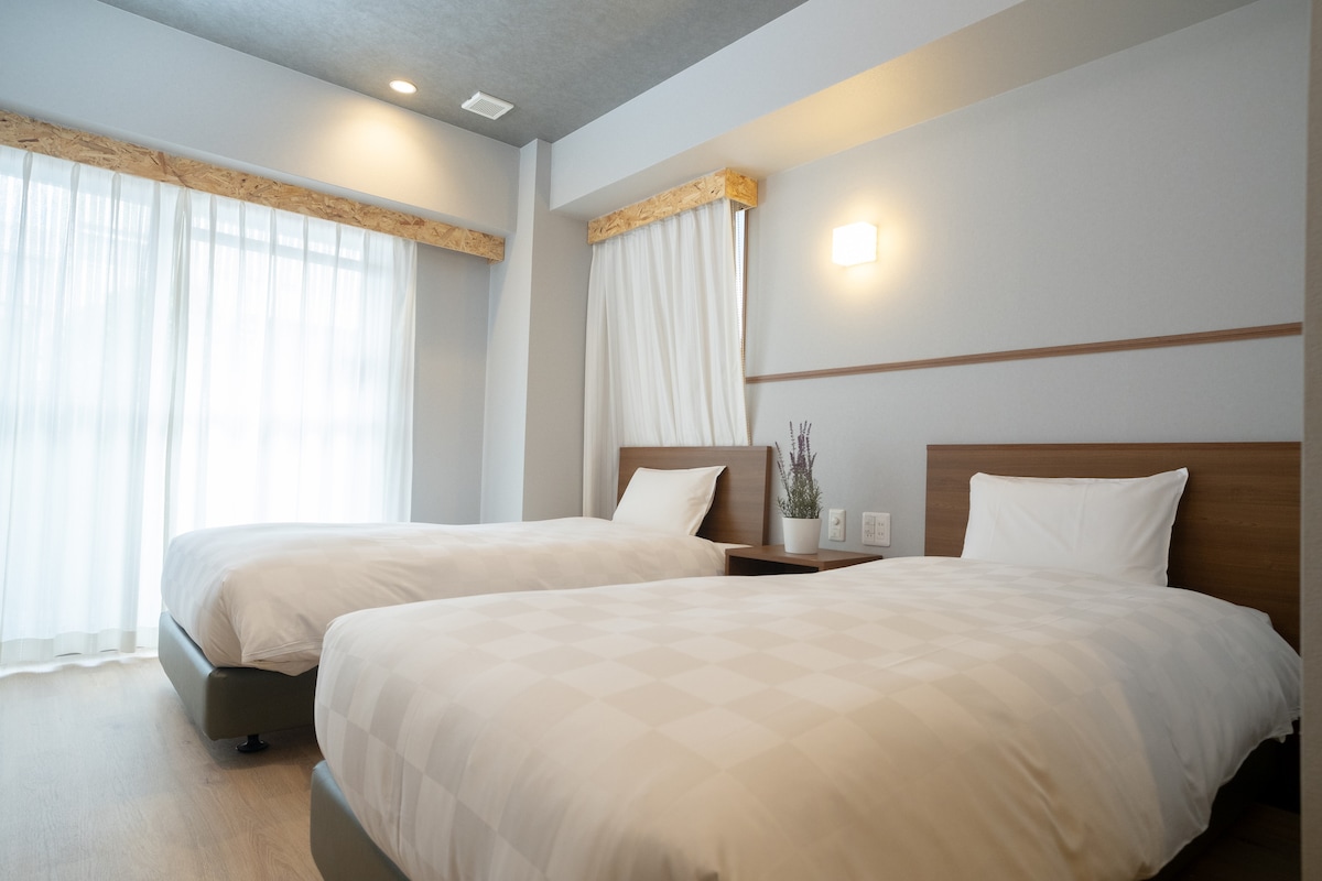 A spacious room features two semi-double beds positioned beside a small bedside table. Large windows allow natural light to illuminate the light-colored walls and wooden accents, while sheer curtains create a soft atmosphere. A simple decorative plant adds a touch of greenery.
