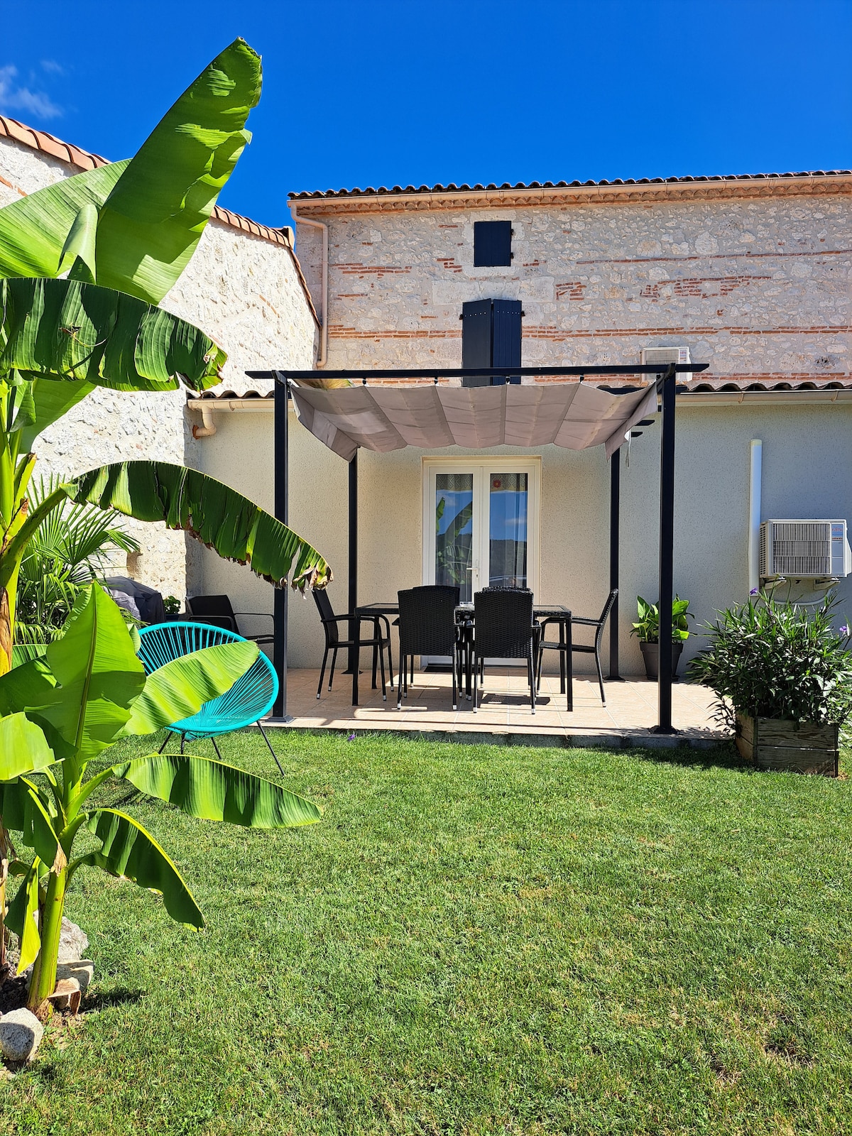 A covered patio is shown with a dining table and chairs, surrounded by lush greenery and a well-maintained lawn. A vibrant banana plant stands prominently in the foreground, while the stone facade of the house provides a rustic backdrop. Natural light enriches the scene.