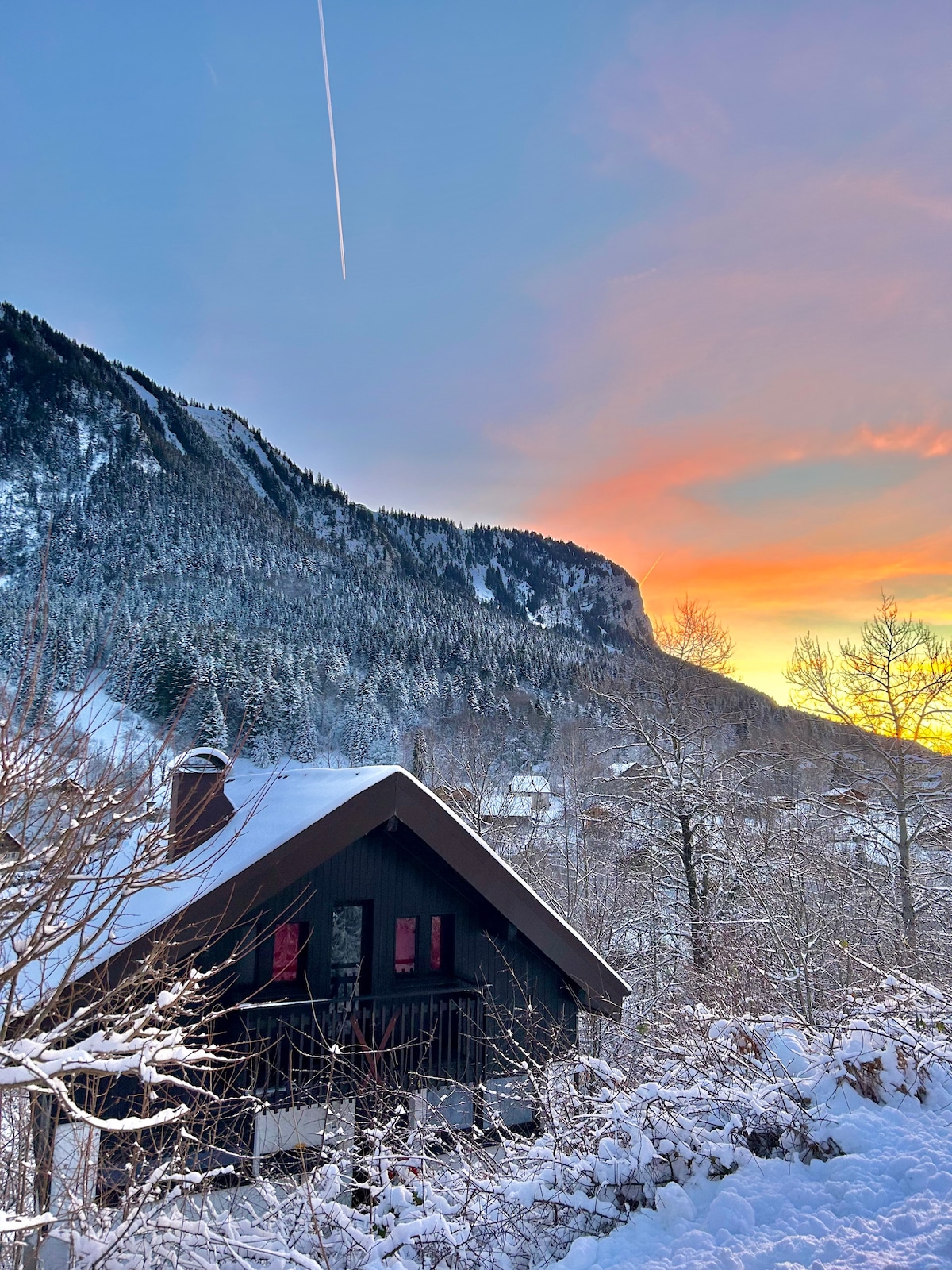 A chalet nestled in a snowy landscape is framed by majestic mountains under a colorful sunset. Snow blankets the ground and trees, while the chalet's dark wooden exterior contrasts with the soft hues of the sky.