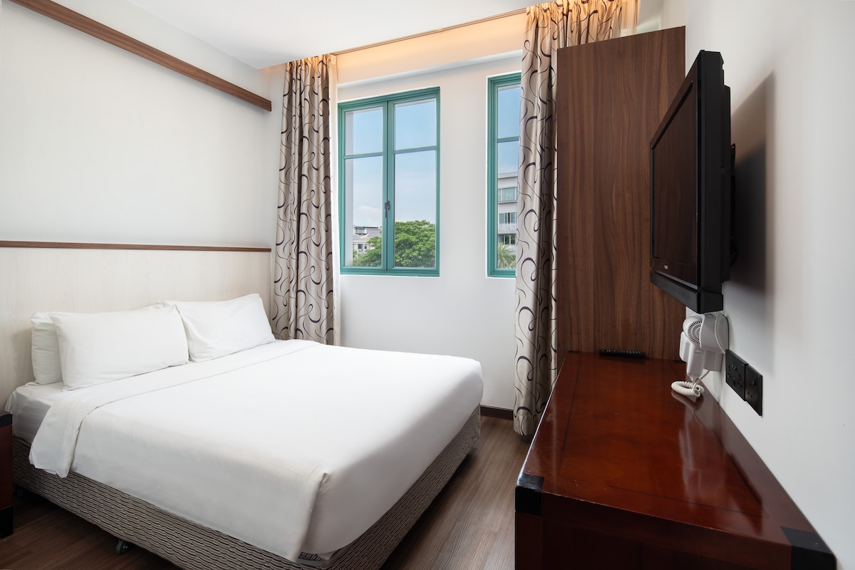 A well-lit guest room is furnished with a spacious bed dressed in crisp white linens. Large windows provide natural light and feature light-colored curtains. A wooden desk with a television is positioned against the wall, contributing to the room's modern design.