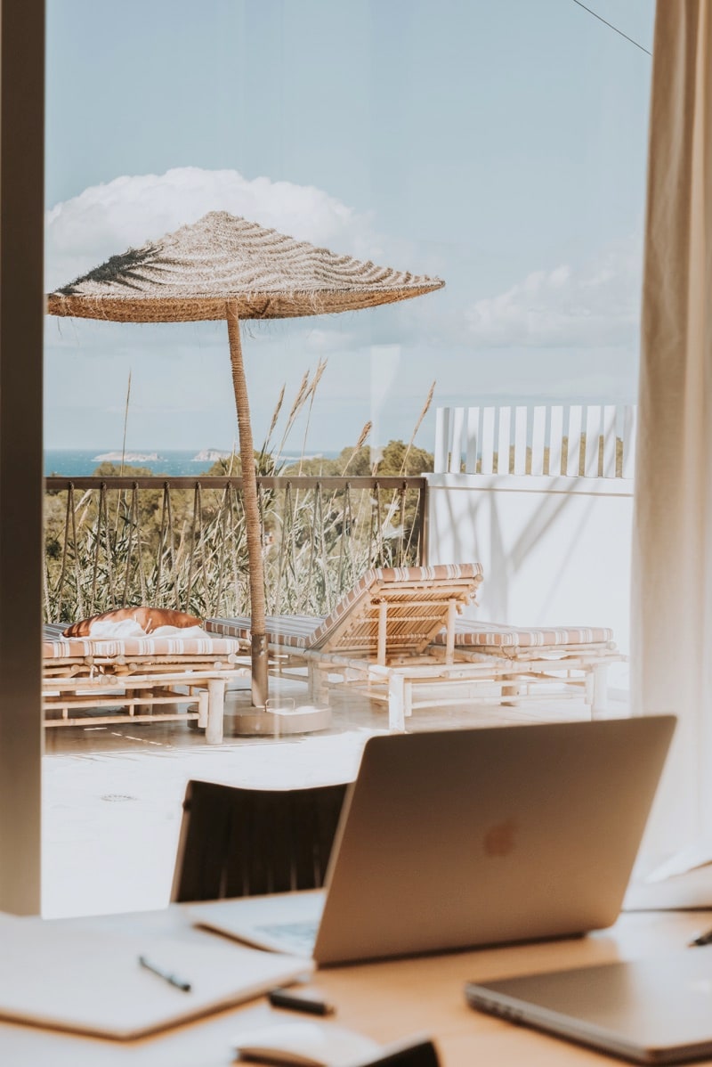 An outdoor terrace showcases a sun umbrella providing shade over a lounge area with wooden seating. Coastal views are visible in the background, complemented by natural landscaping. A workspace featuring a laptop and stationery is positioned near the window, offering a connection to the serene outdoor scenery.