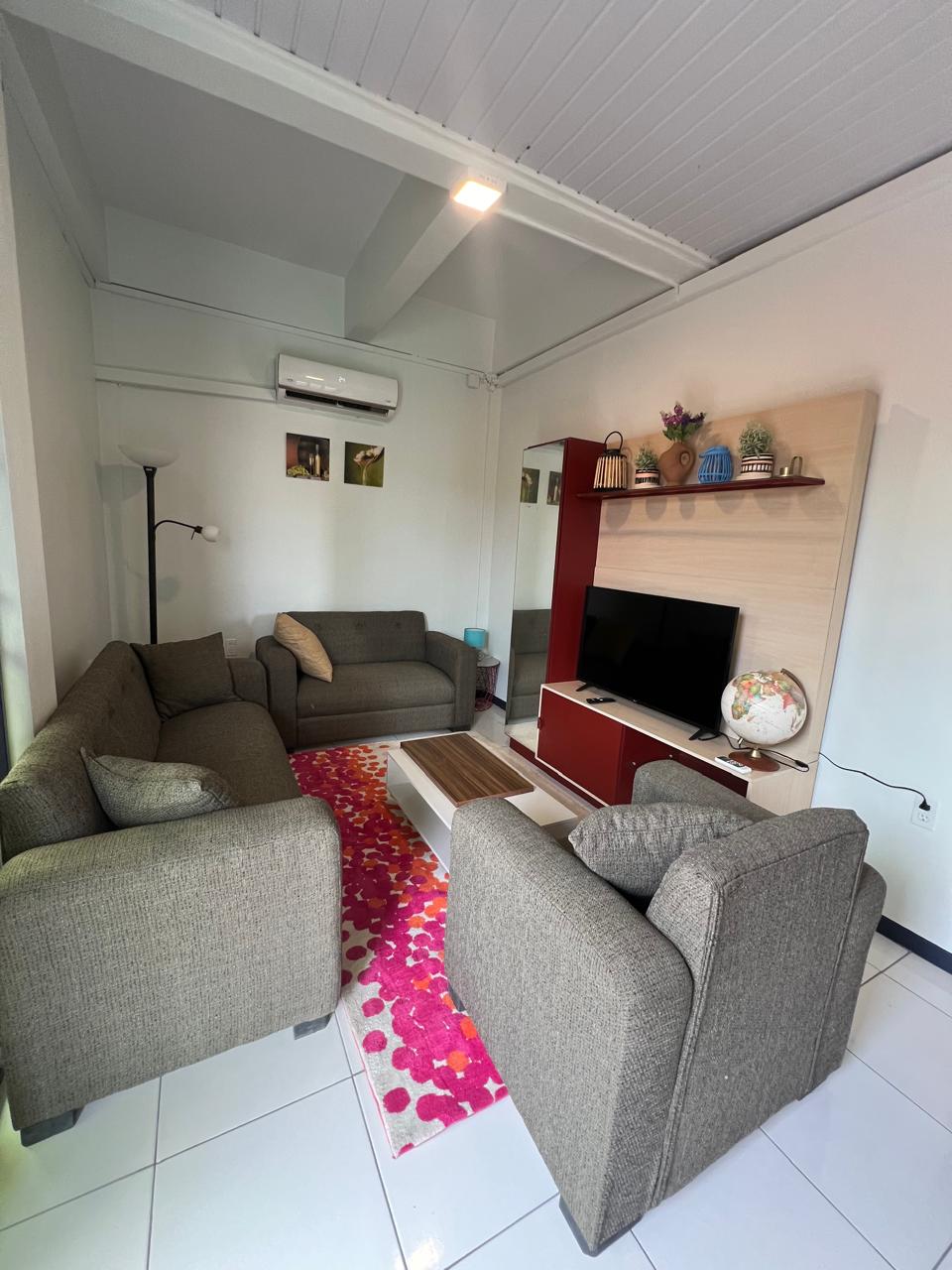 A welcoming living area is furnished with two comfortable sofas and an armchair, arranged around a coffee table. A television is mounted on a light-colored wall unit. Bright patterns adorn the rug, and natural light enters through a nearby window.