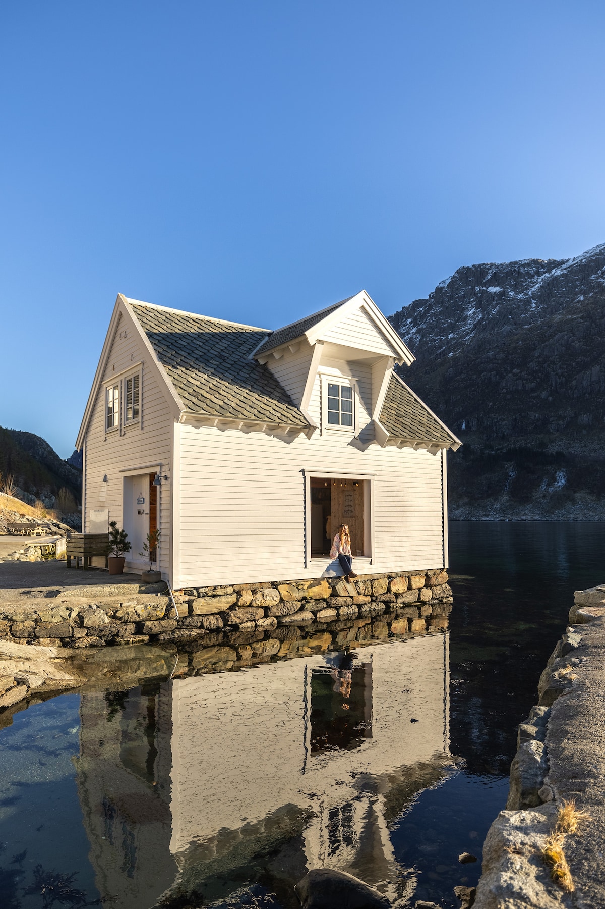 A charming traditional seahouse stands on a stone foundation by the water, reflecting softly in the calm fjord. The exterior features white wooden walls and a steeply pitched roof, framed by a stunning mountain backdrop under a clear blue sky.