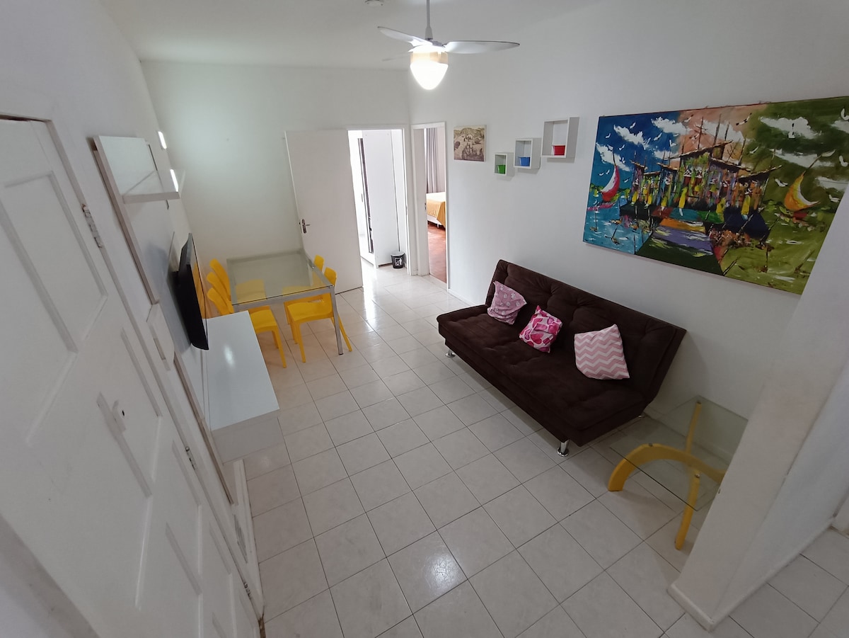 A spacious living area is defined by white walls and tiled flooring. A dark brown sofa bed is positioned against one wall, accompanied by colorful throw pillows. A dining table with yellow chairs is visible, along with a bright painting that adds a touch of color.