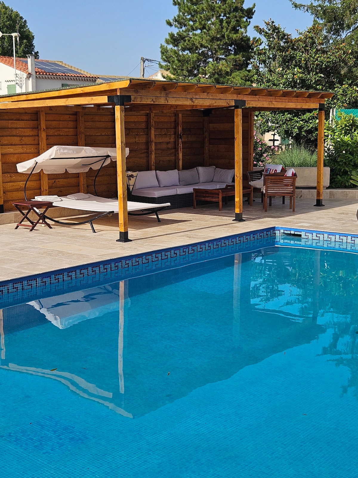 A spacious pool area features a clear blue pool surrounded by tiled flooring. Nearby, a wooden pergola provides shade over a cozy seating arrangement, complemented by wooden lounge chairs. Lush greenery adds a natural touch, enhancing the inviting outdoor space.
