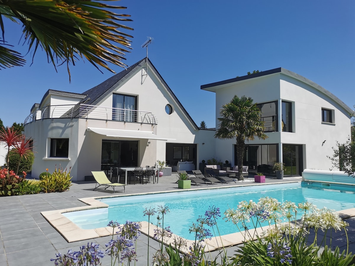 A modern property is showcased with a private pool surrounded by lush greenery. Sun loungers and a dining table are visible near the water, creating an inviting space for relaxation. The architecture features large windows and a stylish design, complemented by vibrant flowers in the foreground.