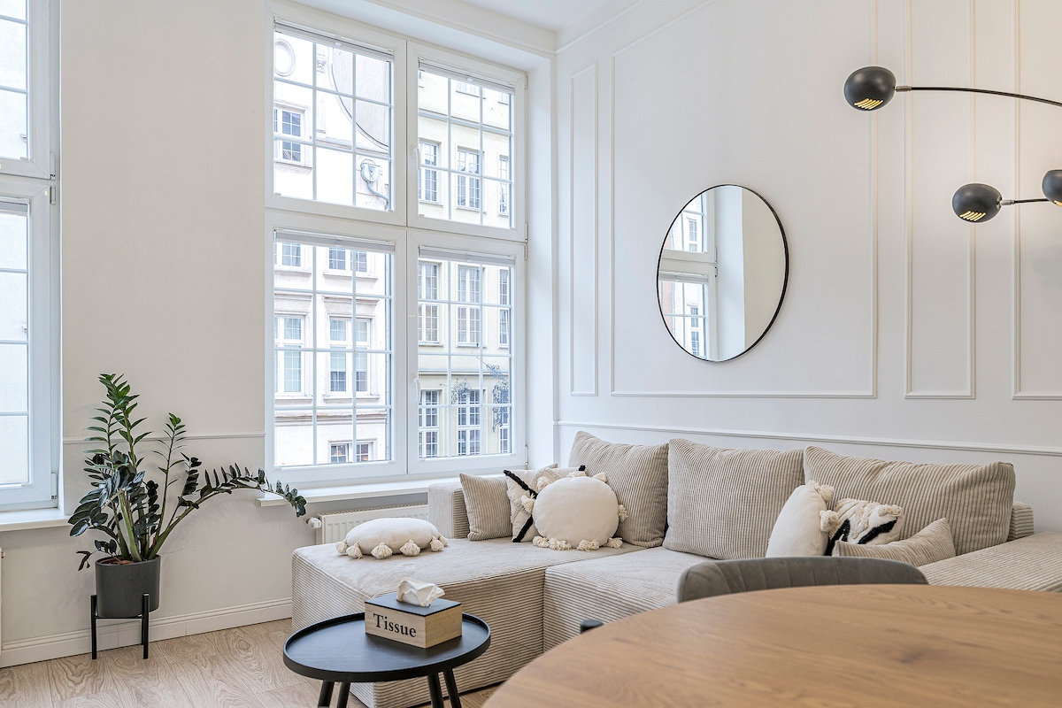 A bright living area is presented, featuring a comfortable corner sofa adorned with various decorative pillows. A round mirror reflects natural light from the large windows. A plant adds a touch of greenery, while a wooden dining table invites guests to gather.