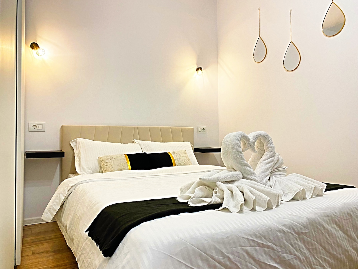 A well-lit bedroom features a double bed dressed in crisp white linens and complemented by a black and gold accent throw. Two wall-mounted lights are positioned beside the bed, and three decorative mirrors hang above it, reflecting the soft light in the space.