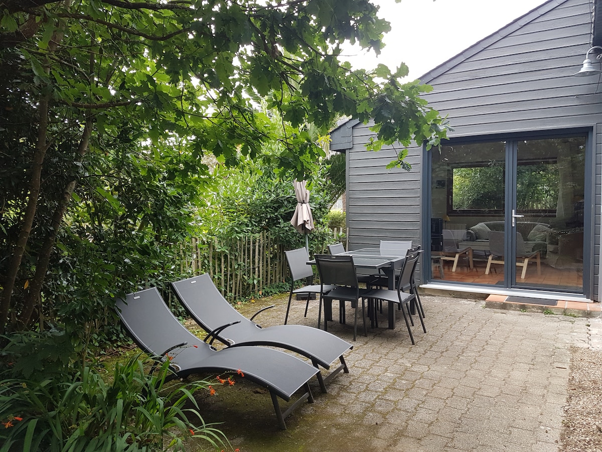An outdoor area is presented, featuring two lounge chairs and a dining table surrounded by greenery. A patio umbrella offers shade, and the grey exterior of the building is visible through large glass doors. Lush plants enhance the tranquil environment.