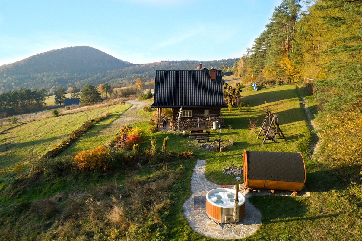 An aerial view of a rustic wooden cottage is presented, surrounded by lush greenery and rolling hills. A wooden hot tub is situated on stone paving, while a swing set and a gathering area are visible in the foreground, enhancing the connection to nature.