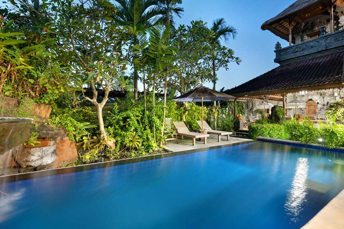 A serene swimming pool is surrounded by lush tropical greenery, featuring comfortable lounge chairs under shaded umbrellas. Soft lighting enhances the tranquil atmosphere as the pool reflects the evening sky, creating a peaceful outdoor retreat.