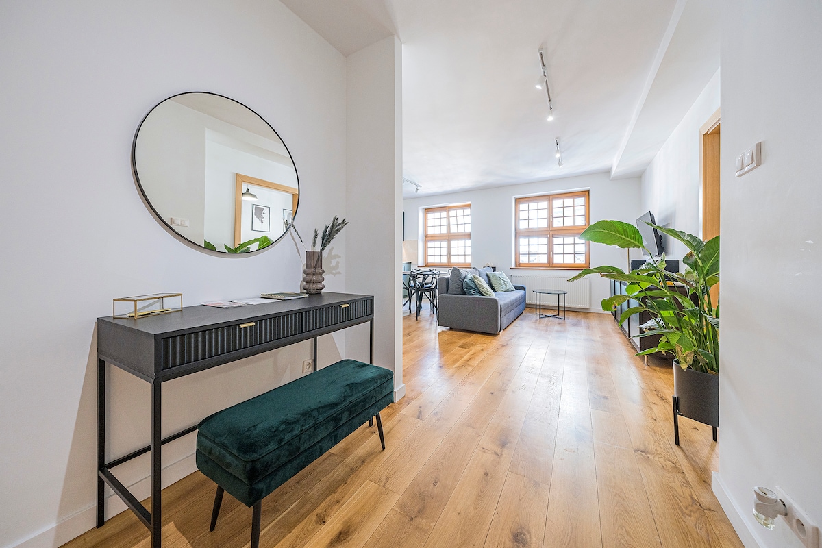 A bright entryway features a chic console table with a dark green bench beside it. Large windows illuminate the space, offering a view of the spacious living area, which is furnished with a modern seating arrangement and decorative plants.