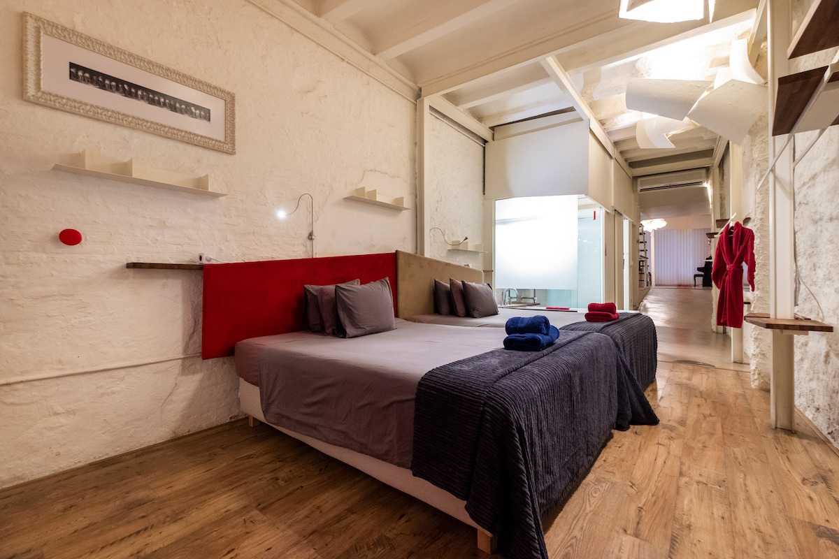 A spacious sleeping area is presented with a large bed featuring a dark blue blanket and layered pillows in grey and red. The room is accented by soft lighting and light-colored walls, creating a calm and contemporary ambiance.