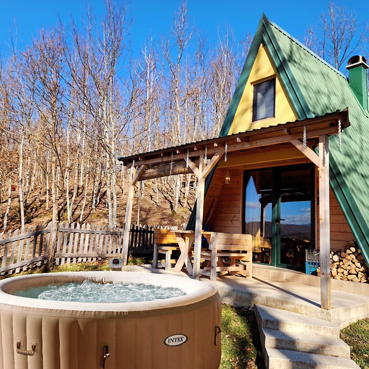 A wooden mountain house in an A-frame design is highlighted with a green roof and yellow accents. In front, a hot tub is positioned under a covered wooden porch, with a view of the surrounding forest and a wooden fence nearby.
