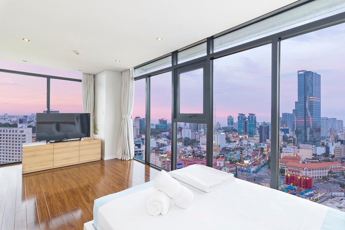 A bright bedroom features a spacious bed with neatly arranged towels and a large window offering panoramic views of the city skyline. Natural light fills the room, enhanced by elegant curtains. A television is positioned on the wooden cabinet, creating a modern living space.