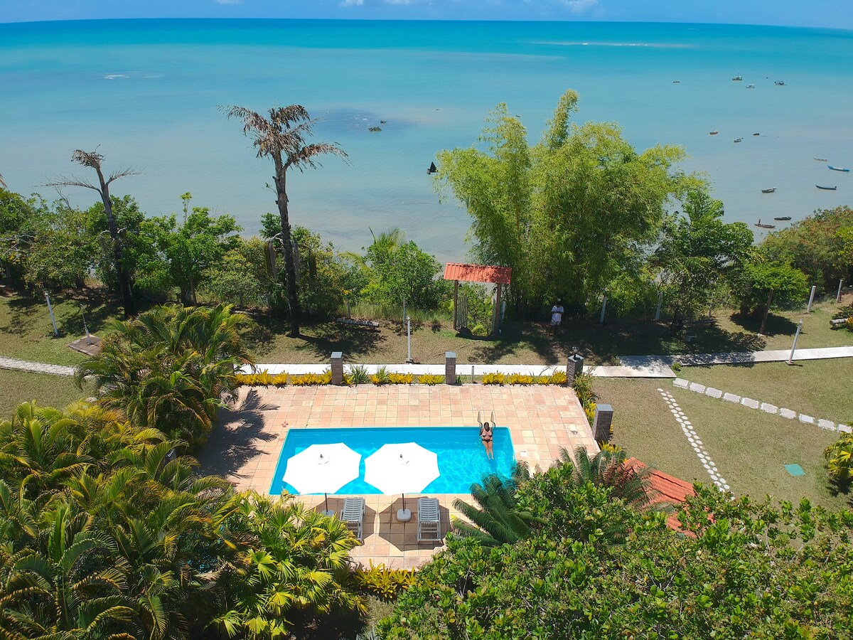 A view from above features a swimming pool surrounded by lush greenery, with two umbrellas providing shade. The pool reflects the clear blue water of the ocean, which is visible in the background, along with a serene shoreline and nearby boats.
