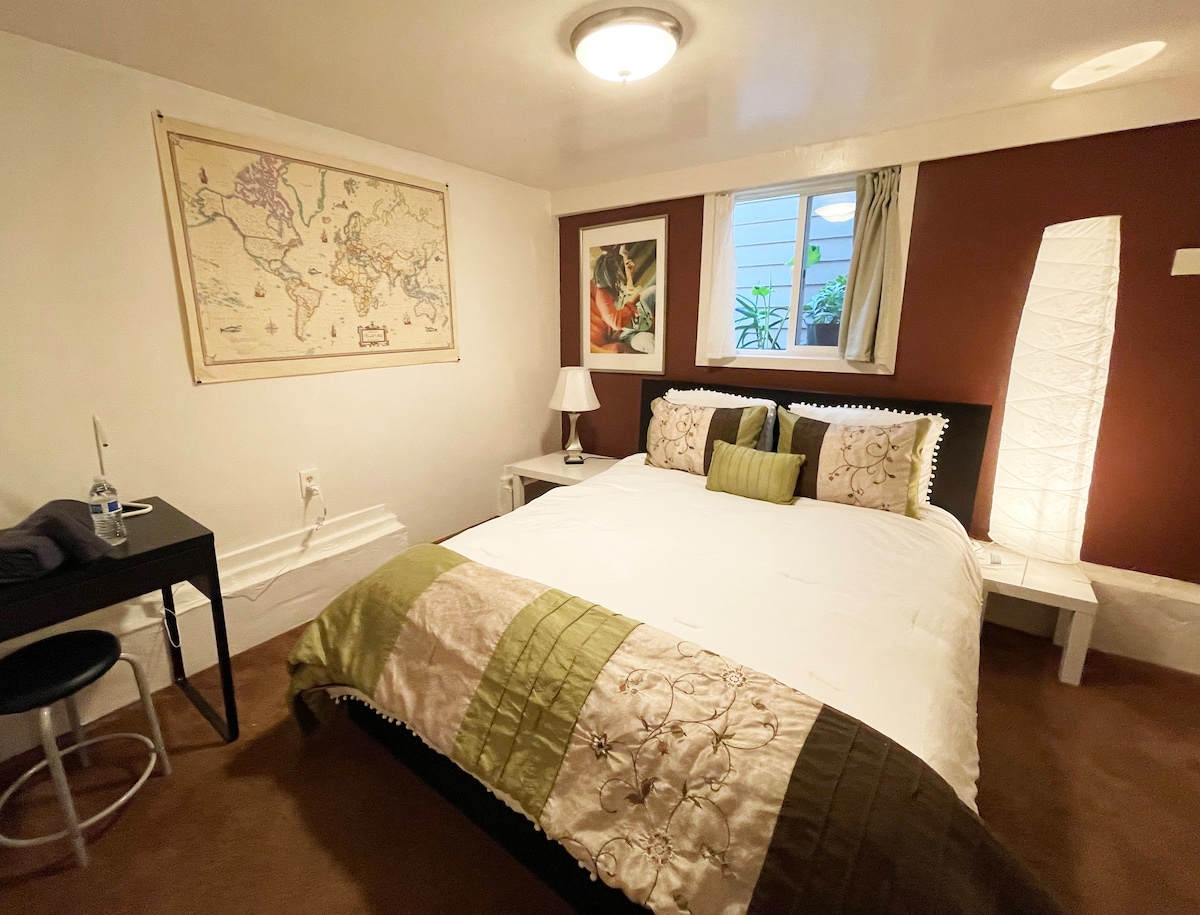 A space featuring a queen size bed with decorative pillows and a textured throw blanket. A light-colored map adorns one wall, while a floor lamp provides soft illumination. Natural light enters through a framed window with a view of greenery, enhancing the cozy atmosphere.