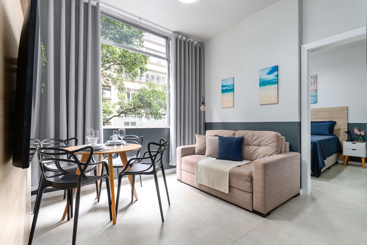 The living area features a comfortable sofa bed and a dining table with four chairs. Large windows allow natural light to fill the space, with views of the greenery outside. Soft grey curtains frame the windows, enhancing the serene ambiance of the room.