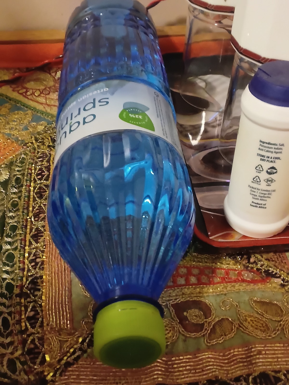 A blue plastic bottle of sparkling water lies on a textured fabric surface. The cap is bright green, contrasting with the bottle's blue hue. Nearby, various containers and a white bottle can be seen, adding to the assortment of items on the table.