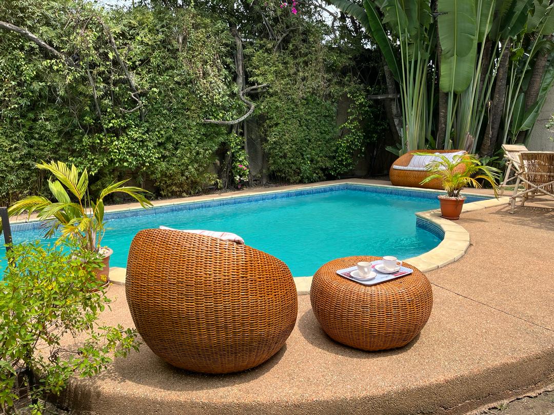 A private pool area features inviting wicker seating arrangements beside a clear blue pool. Lush greenery surrounds the area, providing a serene environment. A small table is set with items, accentuating the space's relaxed atmosphere.