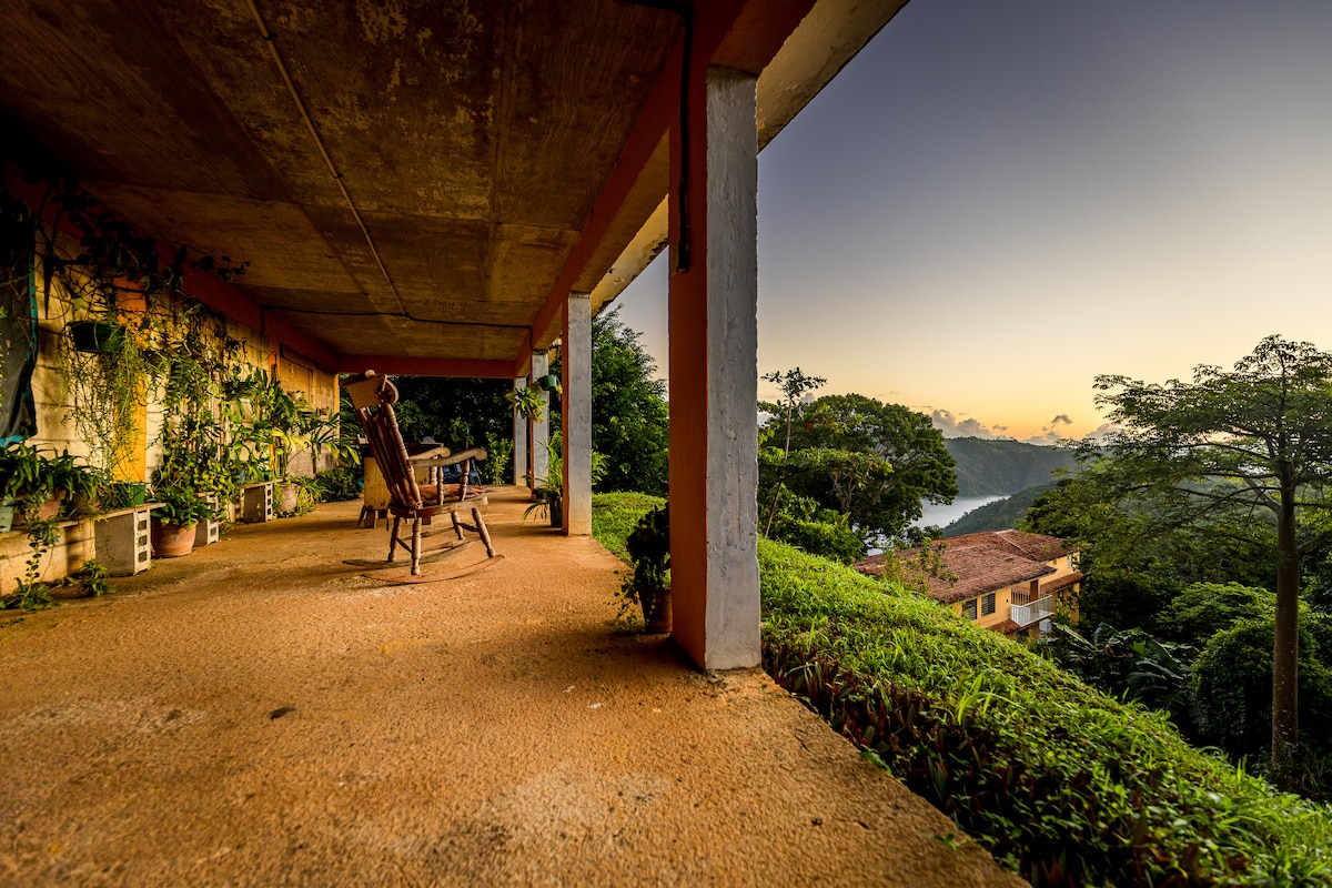 A spacious terrace is seen with comfortable rocking chairs positioned for enjoying the surrounding greenery and the scenic canyon view. Lush plants add a natural element, complementing the tranquil atmosphere as the sun sets in the distance.
