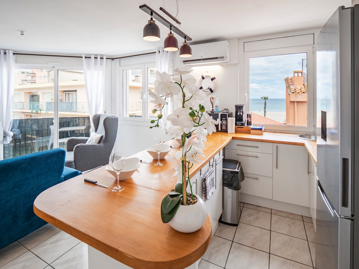 A modern kitchen area features a wooden countertop with bar stools, backed by large windows providing a view of the sea. Fresh orchids add a touch of elegance. Bright light illuminates stainless steel appliances and a spacious layout, creating an inviting cooking space.