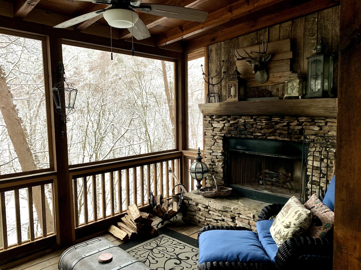A cozy outdoor space featuring a stone fireplace surrounded by wood accents. Comfortable seating with plush blue cushions invites relaxation. A small table sits in front, and stacks of firewood are neatly arranged nearby. Large windows ensure a connection with the serene natural surroundings.