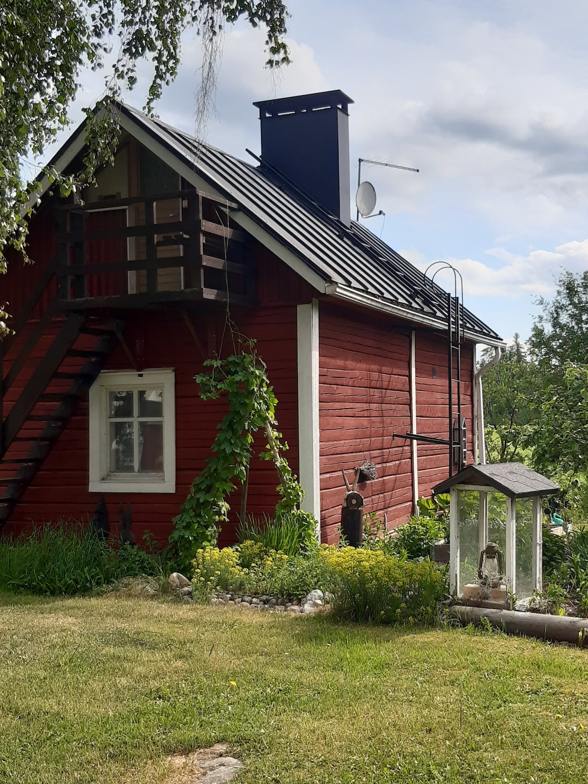 A charming two-story cabin features a red wooden exterior with a black roof and a staircase leading to a small balcony. Lush greenery surrounds the garden area, with a lantern post and decorative stones adding character to the landscape.