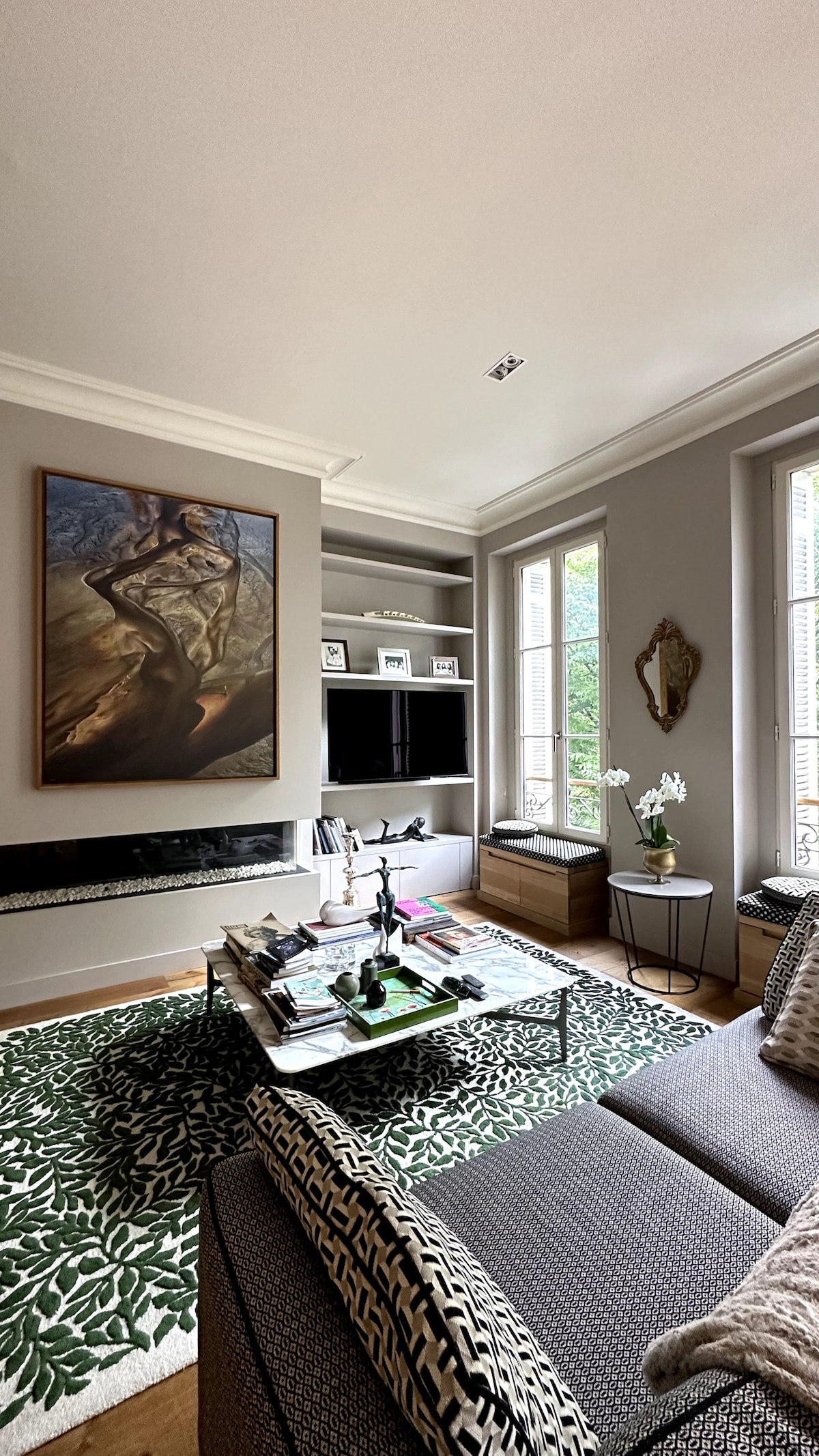 A cozy living area features a modern sofa adorned with textured pillows. A coffee table is set with magazines and decorative items. Two large windows provide natural light, while a piece of art adorns the wall above a minimalist entertainment setup.