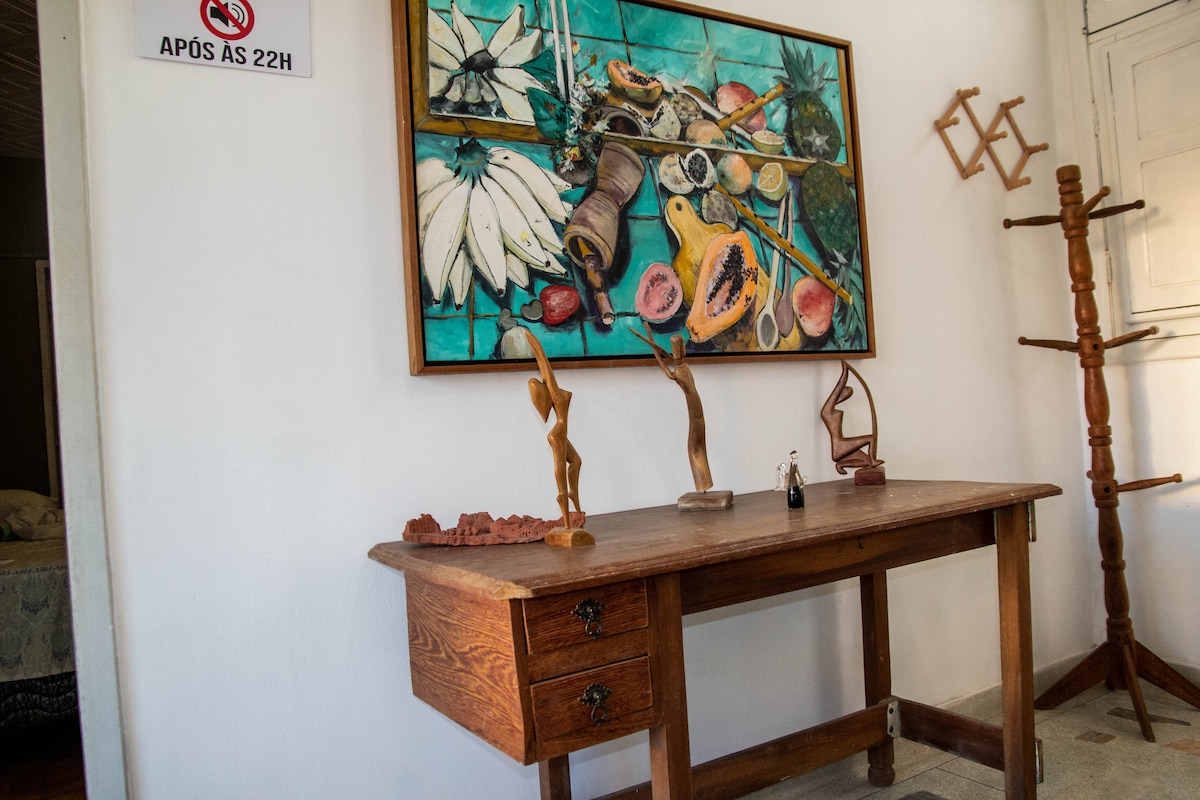 A wooden table is positioned against a white wall, featuring two drawers on the left side. An array of artistic sculptures rests on the table, complemented by a colorful framed painting depicting various fruits. A coat rack stands nearby, adding to the room's functionality.