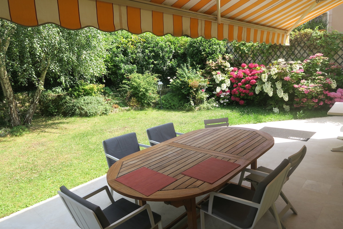 An outdoor dining area features a wooden table surrounded by six chairs, with red place mats on the table. A striped awning provides shade to the space, while lush greenery and vibrant pink flowers create a serene garden atmosphere.