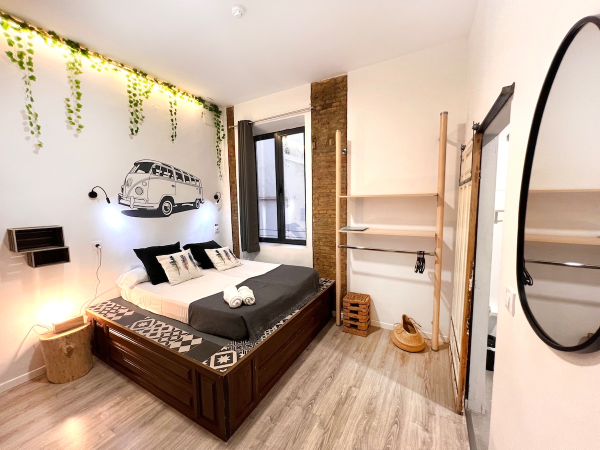 A well-appointed room features a spacious bed adorned with neutral linens and decorative pillows. Natural light enters through a window, while a vintage van mural adds character to the wall. Decorative elements include hanging plants and wooden accents, creating a cozy ambiance.