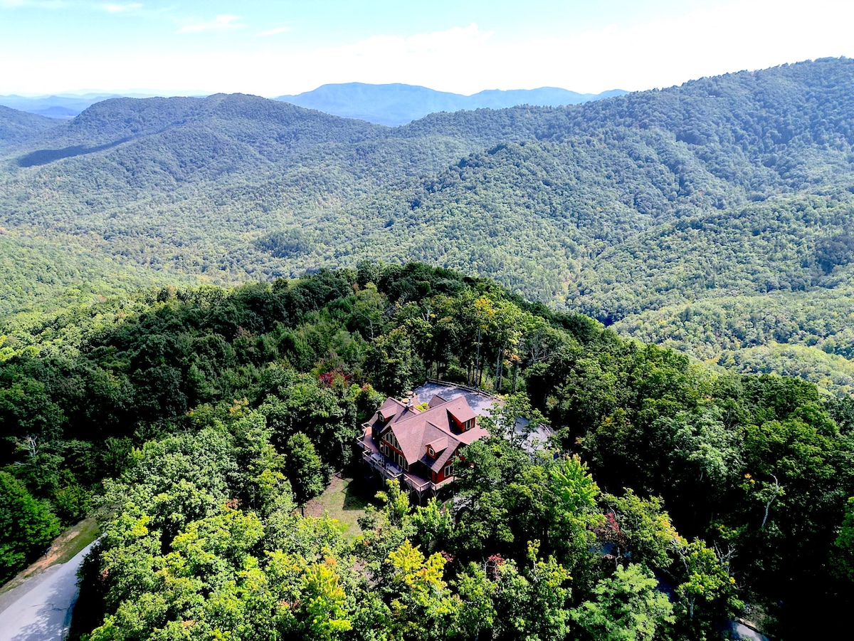 An aerial view captures a luxurious log cabin nestled among a lush landscape of greenery, surrounded by rolling mountains. The expansive deck is visible, highlighting the outdoor space. The distant mountains create a serene backdrop, showcasing the cabin's stunning location.