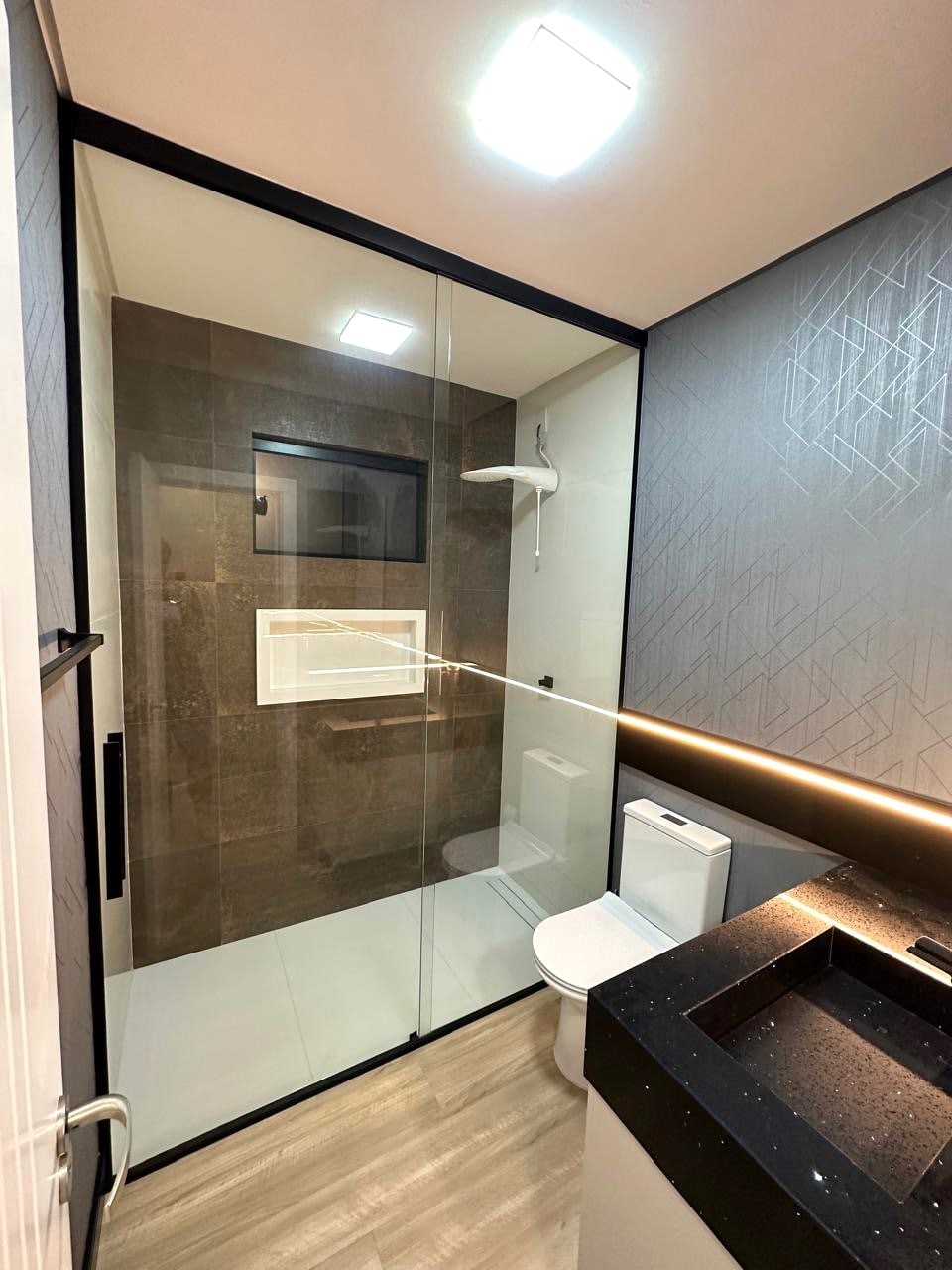 A modern bathroom is showcased with a clear glass shower enclosure, featuring a rainfall showerhead. The room is designed with neutral tones and textured wall panels. A wall-mounted toilet and a dark-colored vanity with a sink emphasize the contemporary aesthetic.
