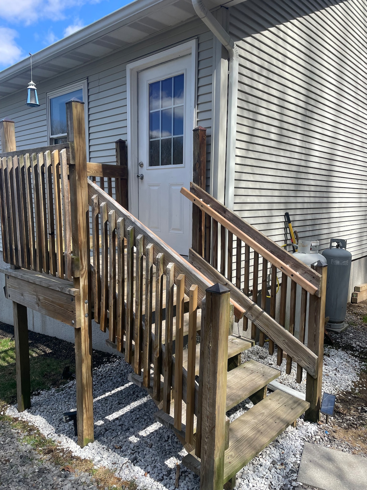 An exterior view of the entrance steps leading up to the apartment, featuring a wooden railing and a door at the top. The stairs are surrounded by gravel and a well-maintained area. Sunlight casts a warm glow on the wooden surface.