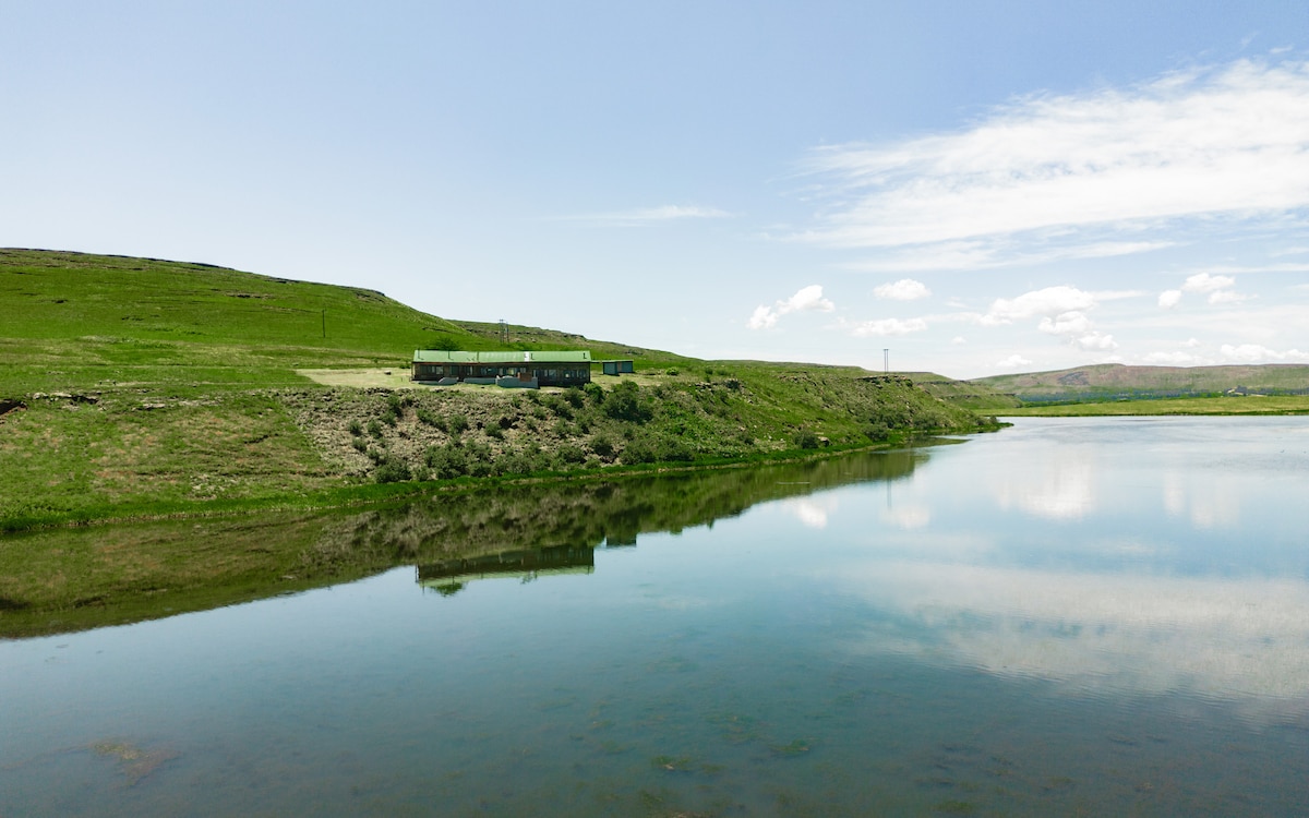 A serene landscape is captured, featuring a calm body of water reflecting the lush green hills and a distant building. The vibrant greenery of the surrounding area complements the clear blue sky dotted with soft clouds.