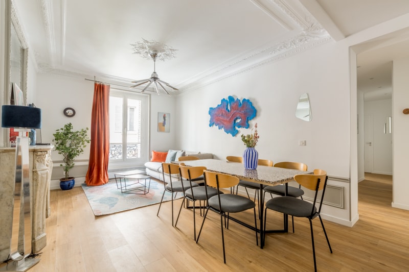 An elegantly furnished living and dining area features a long table surrounded by six chairs. A colorful wall art piece adds visual interest above a comfortable sofa. Natural light fills the space through large windows, with neutral tones complemented by a vibrant curtain and decorative plants.