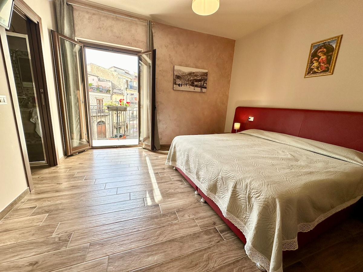 A spacious bedroom is shown, featuring a large red bed with a textured white bedspread. Natural light enters through the open French doors, leading to a balcony. A wall-mounted artwork and light wood flooring add subtle detail to the room.