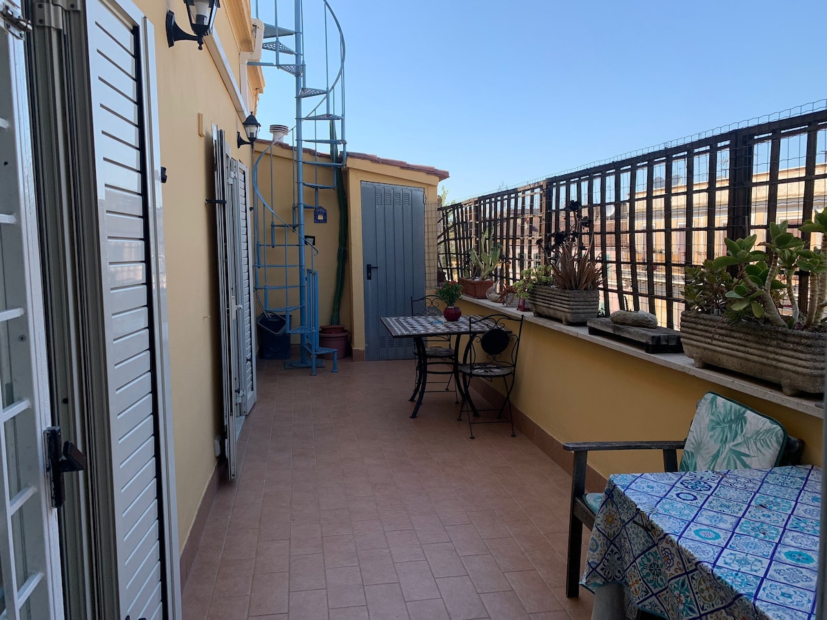 A spacious terrace is depicted, featuring terracotta flooring and a mix of potted plants. A dining table with black metal chairs is set against one wall, while a small, patterned table with a single chair occupies a corner. A spiral staircase is visible, leading to another level.